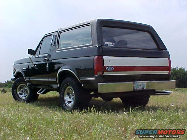 Black Ford Bronco parked in a grassy field, rear view. Has a chrome bumper and red/white stripe on the tailgate