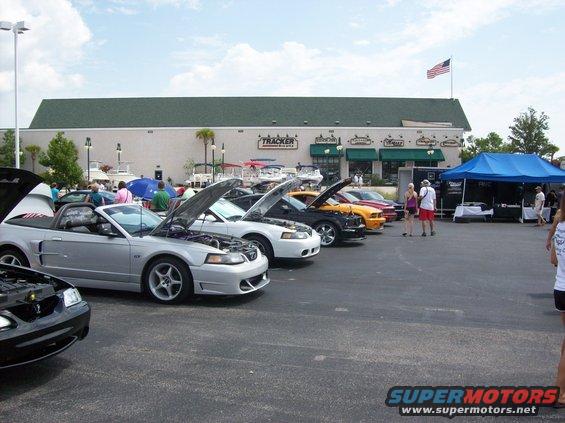 Several Ford Mustangs parked in a lot with hoods open, a building and American flag in background