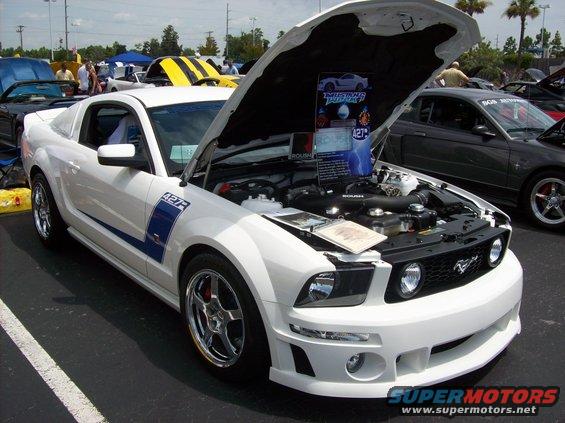 White 2007 Ford Mustang with hood open, showing engine and display board. Another Mustang is parked behind it