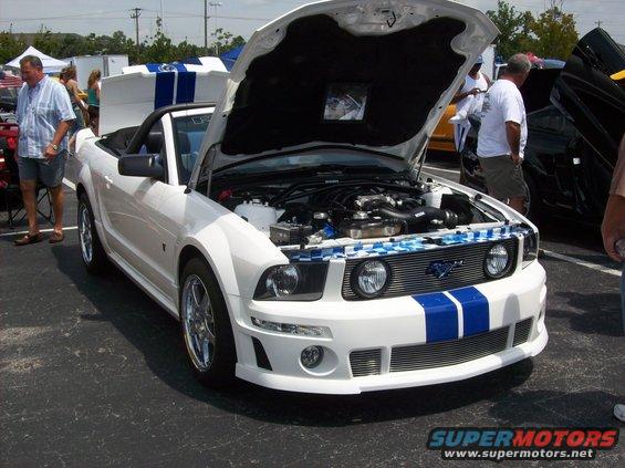 White convertible Ford Mustang with blue stripes, hood open showing engine, parked outdoors