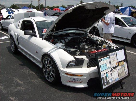 White Ford Mustang with racing stripes, hood open revealing engine, and a display board in front