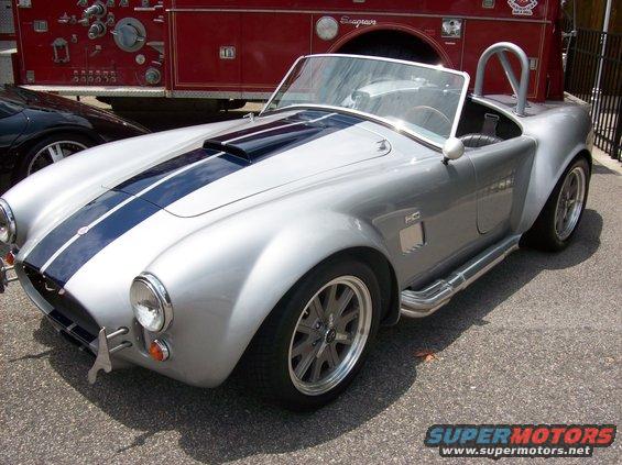 Silver convertible sports car with dual blue racing stripes and side exhaust pipes