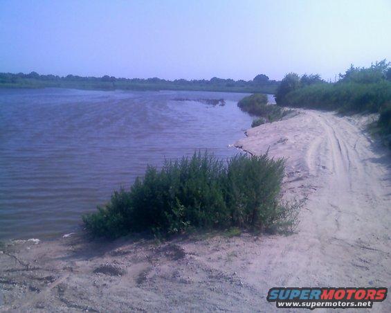 Dirt road curves along a calm body of water with green bushes and trees in the background