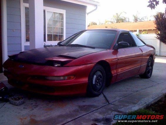 Red coupe with a black hood parked on a driveway, front-left quarter view
