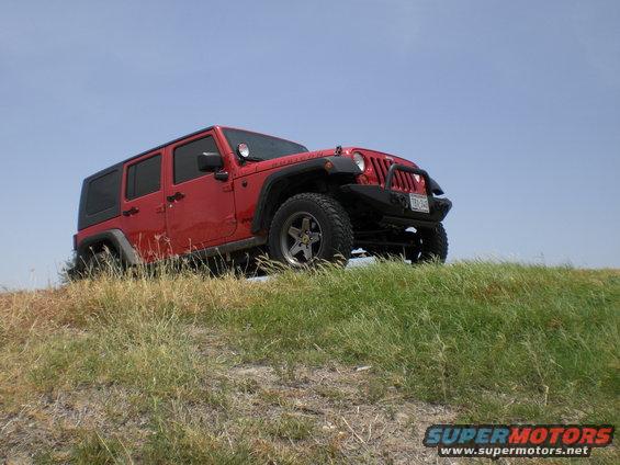 red four-door Jeep Wrangler parked on a grassy hill under a clear blue sky. License plate is visible