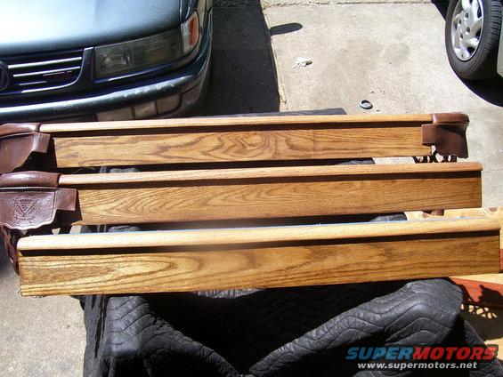 Three wooden pool table rails with brown leather pockets sit on a black quilted cover in front of a dark green car
