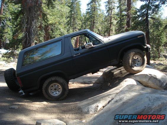 Black Ford Bronco climbing over rocks on a dirt trail, trees in background