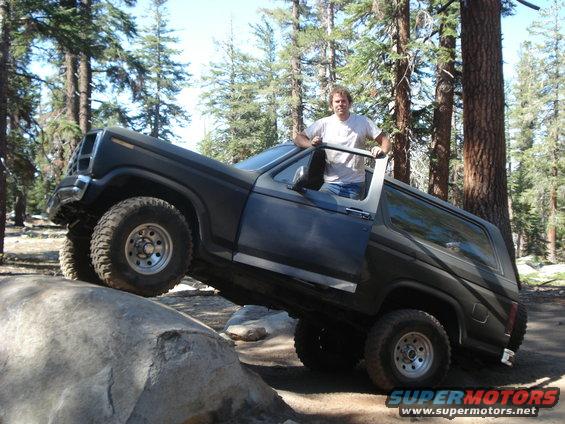 Dark SUV with off-road tires climbing a large rock, driver standing in the open door