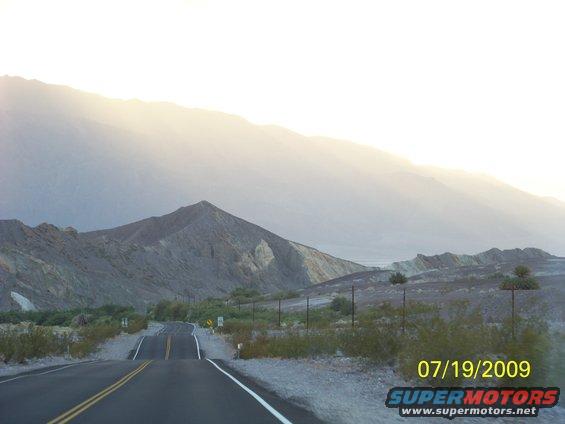 Desert road leading towards mountains under a hazy sky, with a date stamp '07/19/2009' and 'SUPERMOTORS' logo