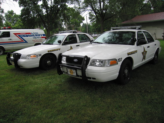 Two white Ford Crown Victoria police cars with push bars parked on grass, one with number 136, the other with 107. A white sheriff van is behind them