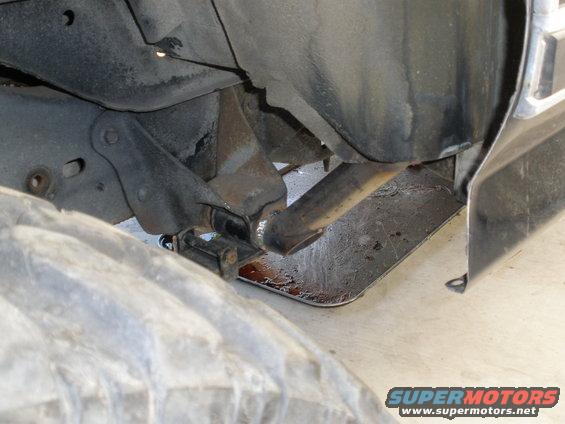 Undercarriage view showing suspension components, exhaust pipe, and a large tire. Some fluid is visible on the ground