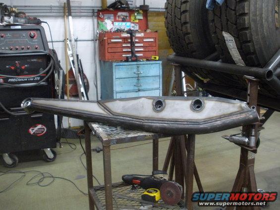 Custom fabricated suspension arm with two mounting holes, resting on a metal table in a workshop. Welding equipment and tires are visible in the background