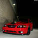 2004 Ford Mustang - A red 2004 Ford Mustang is parked in front of a brick wall at night, its distinctive front grille and black hood stripe.