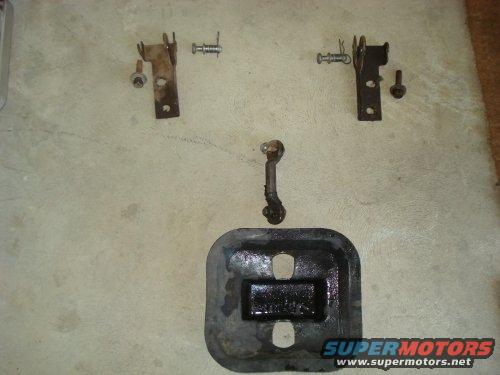 Several metal vehicle parts, including brackets, bolts, and a rubber-coated plate, laid out on a concrete surface