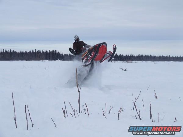 Snowmobile airborne in a snowy field with trees in the background