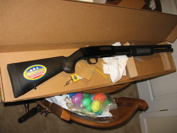 Shotgun with 'Made in USA by Mossberg' sticker on the stock, sitting in a cardboard box with a bag of plastic Easter eggs