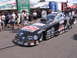 1998 GMC Sierra 1500 - A black dragster with sponsor decals is parked on a paved surface, with people and tents visible behind it.