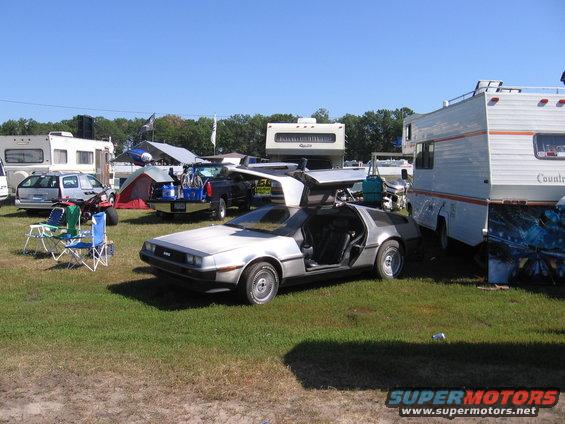 Silver DeLorean DMC-12 with gullwing doors open, parked on grass next to RVs