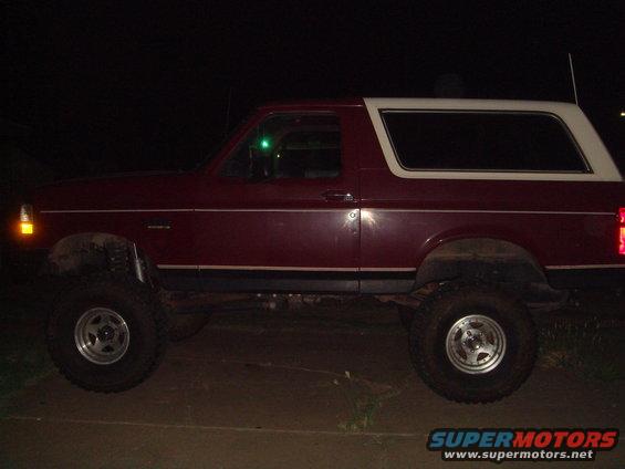 Maroon 1992 Ford Bronco with white top, lifted suspension, and aftermarket wheels, seen from the side at night
