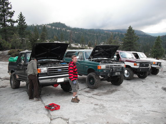 Four Ford trucks and a Bronco parked on rocks, hoods up, with people working on them. Mountains and trees in background