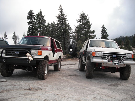Two lifted Ford Broncos parked on rocks, one red and white, the other silver and gray, with trees in the background