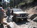 1991 Ford Bronco - Two Ford Broncos navigate a challenging, rocky off-road trail in a wooded area.