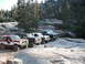 1991 Ford Bronco - A lineup of four off-road SUVs, including a 1991 Ford Bronco, parked on a rocky bank beside a flowing stream.