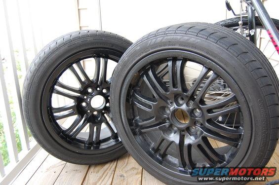 Two black alloy wheels and tires sit on a wooden deck. A bicycle is visible in the background