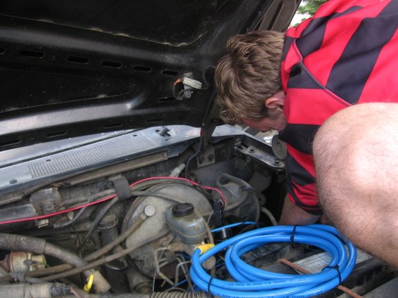 1989 Ford Bronco engine bay with a person in a red and black striped shirt working on the engine. Blue tubing is coiled in the foreground