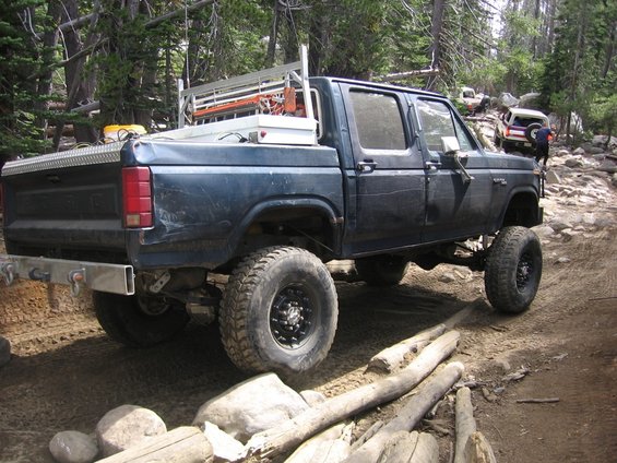 Dark blue Ford F-Series pickup truck with a toolbox and rack in the bed, lifted suspension, and off-road tires, driving on a rocky trail