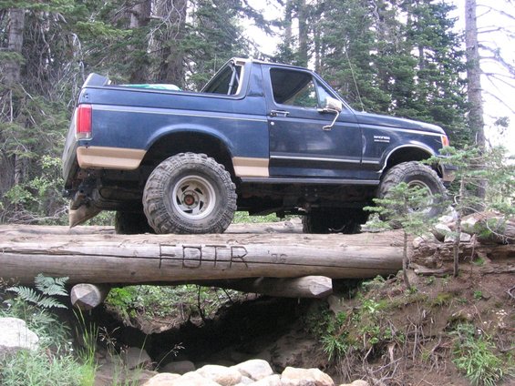 Blue 1989 Ford Bronco with tan trim driving over a log bridge in a forest