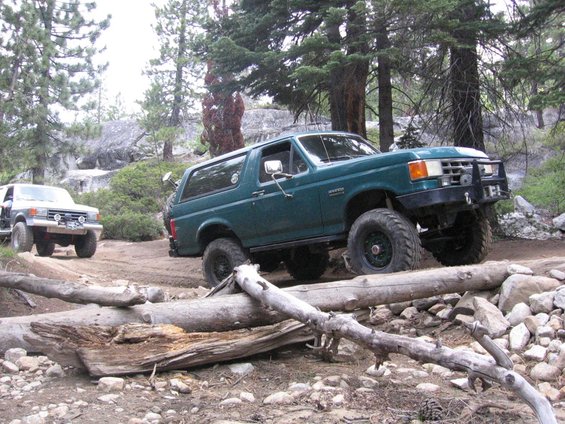 Green Ford Bronco driving over logs on a dirt trail, another SUV behind it