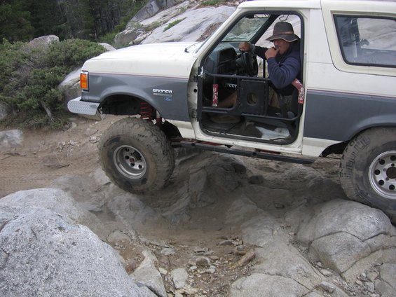 White and grey 1989 Ford Bronco off-roading over rocks, driver wearing a hat