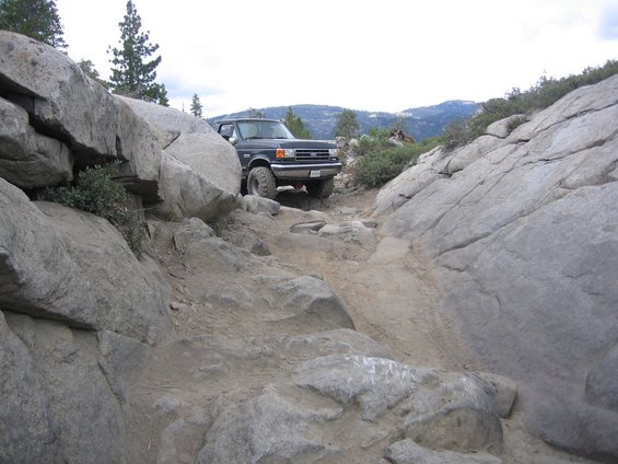 1989 Ford Bronco navigating a steep, rocky off-road trail with large boulders on either side