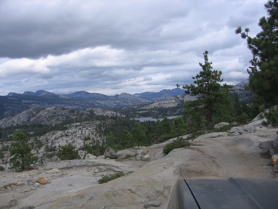 Dark gray vehicle hood in foreground, overlooking a vast mountain range with a lake in the distance under a cloudy sky