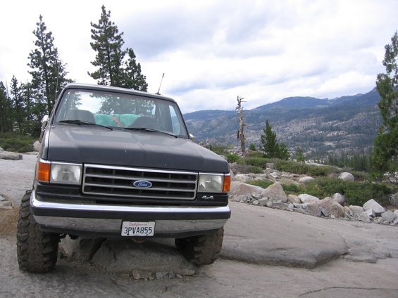 Dark gray Ford Bronco with off-road tires parked on rocks, mountains in background. License plate 3PVA855