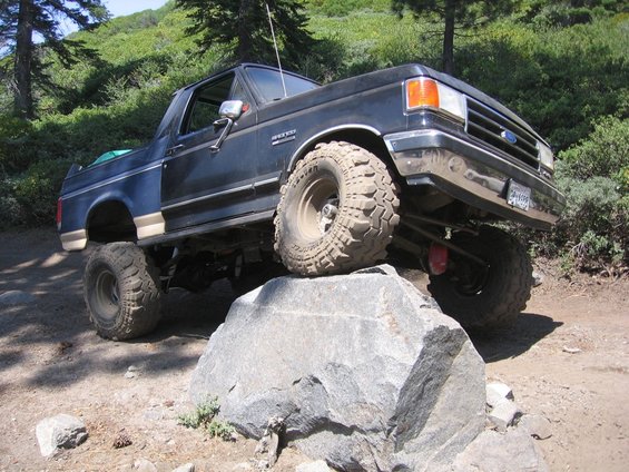 Black 1989 Ford Bronco with lifted suspension and large tires climbing over a rock on a dirt trail