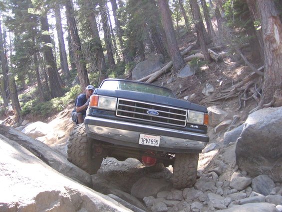 Front view of a dark blue Ford Bronco with large tires navigating a rocky trail. License plate reads 3PVA855