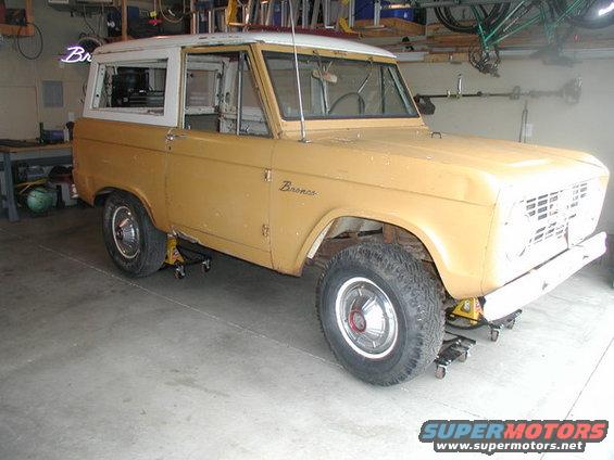Yellow 1966 Ford Bronco on jack stands in a garage. White hardtop. Front grille is removed