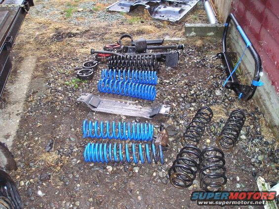 Several sets of blue and black coil springs, suspension components, and a metal chassis part laid out on gravel near a wooden building