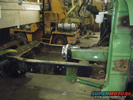 Green vehicle body section with exposed frame and suspension components. Forklifts and toolboxes in background