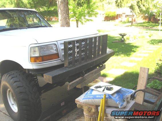 White Ford truck front end, showing headlights, grille, and custom metal bumper with mesh insert. A large tire is visible