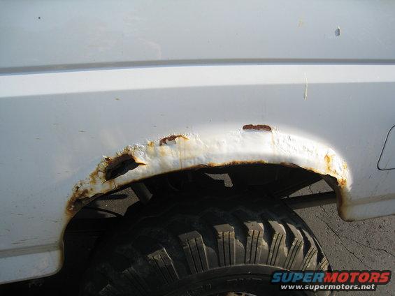 White SUV's heavily rusted rear wheel well with exposed metal and tire visible