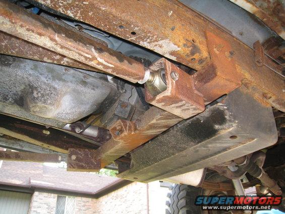 Underside of a rusty vehicle frame with suspension components visible. A SuperMotors logo is in the corner