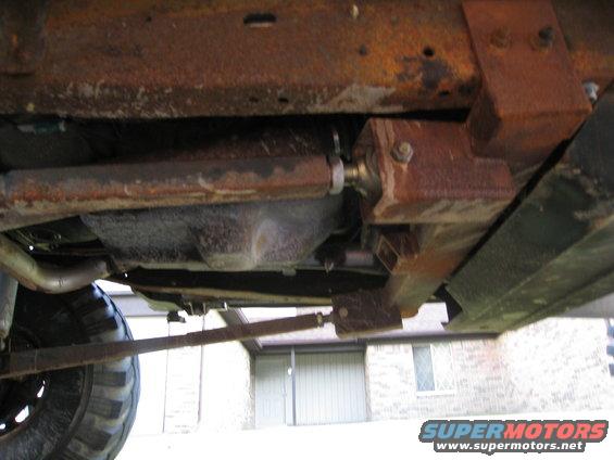 Undercarriage of a heavily rusted vehicle, showing suspension components and exhaust pipe. A tire is partially visible