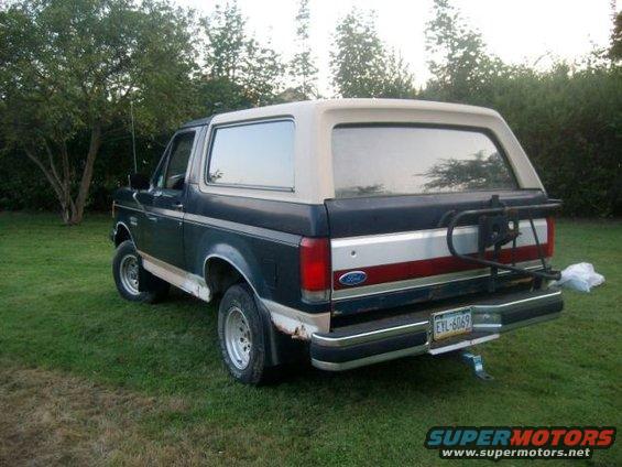Rear view of a dark blue Ford Bronco with a tan top, parked on grass. It has a spare tire carrier on the back and a license plate that reads EYL-6069