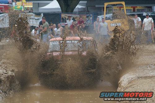Red SUV splashing through mud pit with a yellow tractor in the background