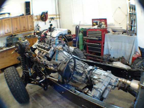 Vehicle chassis with engine and transmission assembly in a workshop. Front suspension and tire visible
