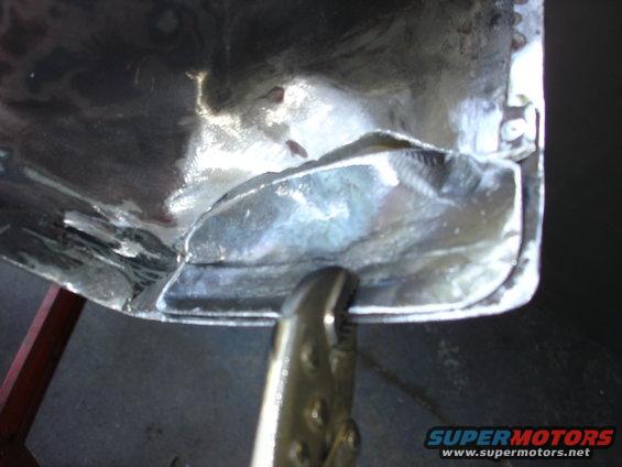 Metal panel with visible welding seams and a clamp holding a section. The panel is shiny and appears to be undergoing repair