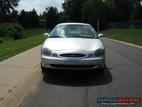Silver sedan parked on asphalt road, front view. The car has oval headlights and a grille with a Ford emblem